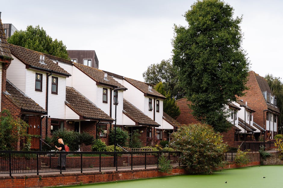 An outdoor view of a residential housing area featuring a row of modern, neatly maintained houses with white painted exterior walls and brown tiled roofs. The scene includes a well-kept garden with lu