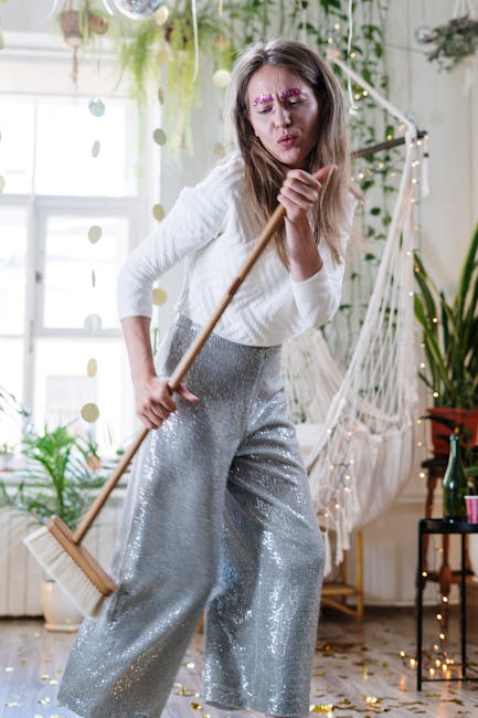 A young woman with long hair, wearing a white sweater and shiny silver trousers, is holding a broom and making a playful face in a bright, well-lit room filled with plants. The room has large windows,