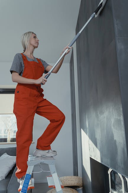 A woman with blonde hair wearing grey and orange cleaning overalls stands on a small step ladder, cleaning a high wall or ceiling with a long-handled duster or cleaning tool. The room has a modern decor with a large black panel or surface and natural lighting coming through a window in the background. The floor appears to be carpeted, and there is a beige cushion or mat on the floor nearby. This image illustrates surface cleaning, dust removal, and maintenance activities related to domestic or commercial cleaning, consistent with the Harringay Ladder carpet cleaning guide for period homes, helping to maintain hygiene and cleanliness, with professional service provided by Cleaners Harringay.