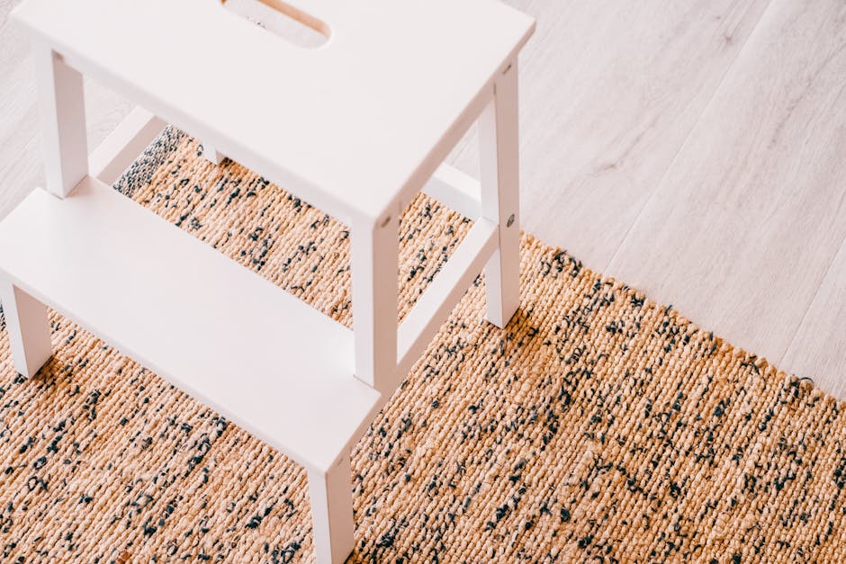 A vacuum cleaner with a black body and a long, slim black tube attachment is being used to clean a patterned woven rug with floral motifs in shades of beige, green, and orange. The cleaning process appears thorough, with a focus on the surface fibers of the rug, which is laid out on a hard floor. Soft sunlight illuminates the scene, highlighting the clean, dust-free appearance of the rug in a well-lit room. In the background, a person, partly visible, is standing nearby wearing casual clothing and sneakers. The image demonstrates surface cleaning within a residential setting, aligning with professional domestic cleaning services provided by Cleaners Harringay, as referenced in the Harringay Ladder carpet cleaning guide for period homes.