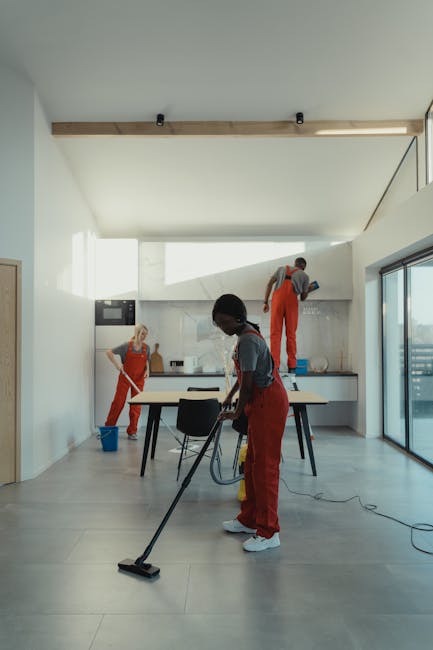 A team of three professional cleaners wearing red aprons and grey shirts is performing a thorough cleaning in a modern, bright kitchen with large glass sliding doors. One cleaner in the foreground is vacuuming the light-colored wooden or ceramic tile floor, focusing on surface cleaning and dust removal. In the background, another cleaner is mopping or wiping down the white kitchen cabinets, counter space, and stainless steel appliances, demonstrating deep cleaning and sanitisation processes. The third cleaner is standing on a step ladder, applying cleaning solutions to an upper wall or cabinet area, ensuring comprehensive coverage. The room features minimalist decor with natural light flooding through the windows, highlighting the clean, shiny surfaces and the meticulous work of Cleaners Harringay in maintaining hygiene and surface cleanliness in a domestic setting.
