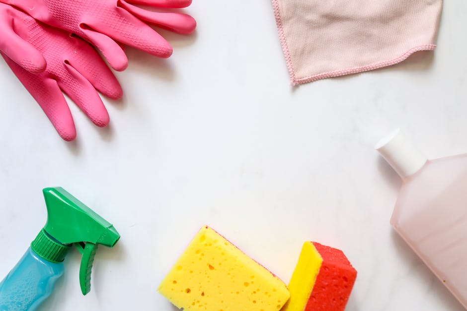 An arrangement of cleaning tools and supplies on a white surface, including a pair of pink rubber gloves, a light pink cloth, a green spray bottle, a yellow and red sponge, and a partially visible white plastic bottle with a pink cap, illustrating materials used for surface cleaning, disinfecting, and maintenance in domestic or commercial environments, as featured on the Cleaners Harringay website in the context of understanding hidden charges in Harringay cleaning quotes.