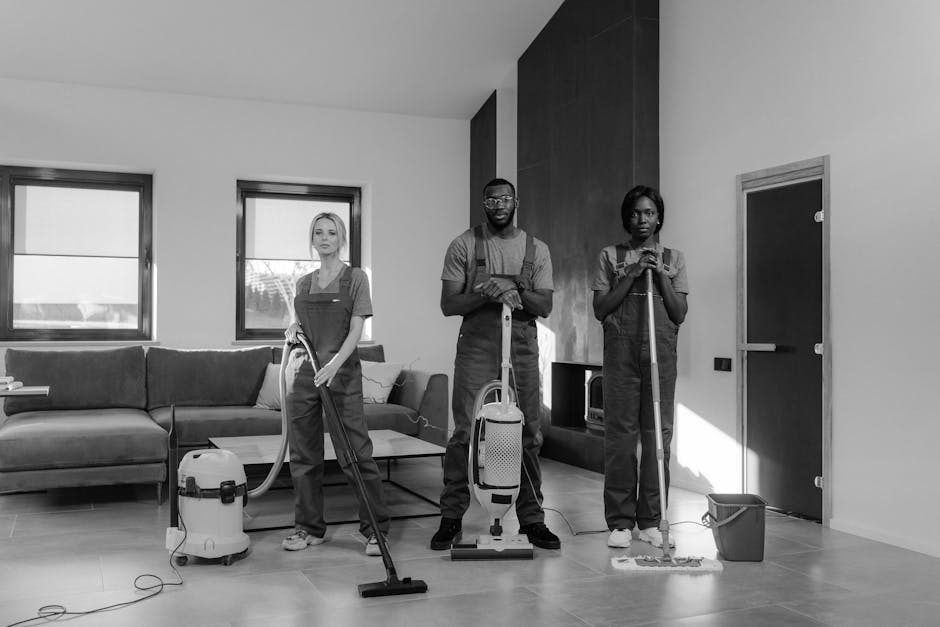A team of three professional cleaners from Cleaners Harringay performing surface cleaning and deep cleaning in a living room with a modern design. The room features a dark-colored sectional sofa, a wo