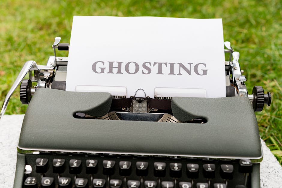 A vintage typewriter outdoors on a stone surface with a paper inserted displaying the word 'GHOSTING' in bold, uppercase letters. The typewriter features a grey-green textured casing, black keys with white lettering, and visible metal mechanisms on either side. The background shows a grassy area, emphasizing the outdoor setting, and the lighting appears natural, highlighting the textures and details of the typewriter and paper. This image illustrates the concept of 'ghosting' related to surface cleaning and maintenance, connecting to the topic of same-day cleaning delays in Harringay, as discussed by Cleaners Harringay.