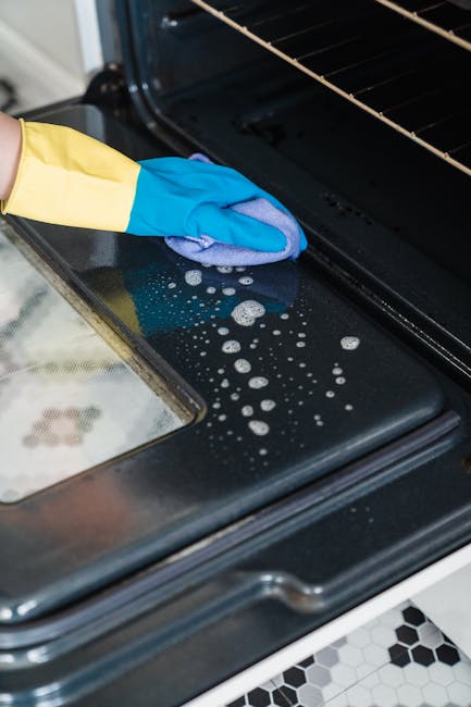 A person wearing yellow protective gloves and a white shirt is performing surface cleaning inside a black oven, using a pink cloth to wipe the oven's glass door, which shows some food splatters. The oven is part of a white freestanding stove with visible control knobs on top, located in a kitchen with patterned black and white floor tiles. The scene emphasizes deep cleaning and hygiene practices typical of domestic cleaning services provided by Cleaners Harringay, focusing on oven maintenance and sanitisation for Harringay homes.
